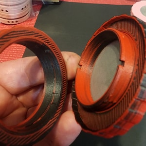 May include: Two red and black 3D printed round objects with a textured surface. One object has a mesh screen inside.