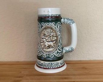 May include: A ceramic beer stein with a white handle and rim, and a decorative base. The stein features a detailed relief of an English Setter dog and hunters. The design includes green and white scrollwork and the words "English Setter".