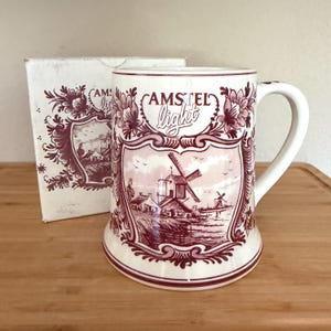 May include: A white ceramic beer mug featuring a maroon-colored transfer print of a Dutch windmill landscape and the text Amstel light. The mug is accompanied by a matching white cardboard box with the same maroon floral and windmill design.
