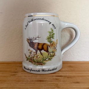 May include: A light gray ceramic beer stein featuring a gold rim and a handle. The front displays a colorful illustration of a stag in a forest setting, with the text 9. Int. Wanderung 1989 and Wanderfreunde Wiesbachtal printed in black.