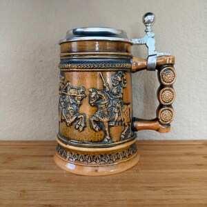May include: A brown ceramic beer stein featuring a relief design of knights on horseback. The stein has a metallic hinged lid and a handle decorated with three circular patterns. The body displays intricate carvings of armored figures.