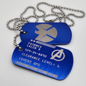 May include: Two blue metal dog tags with a silver chain. The top tag features a silver shield with a star and a wing. The bottom tag has a silver Avengers logo and the text "FAIRRES CALEB D 729-34-8892 CLEARANCE LEVEL: 6 COVERT OPS".