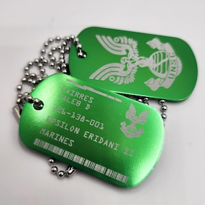 May include: Two green metal dog tags with silver text and chain necklaces. One tag features a winged emblem and the letters "UNSC". The other tag has the name "Fairres Caleb D", a number, and the words "Epsilon Eridani II Marines".