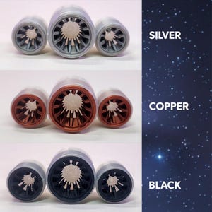 May include: Three bottle stoppers in silver, copper, and black. Each stopper has a cylindrical design with a detailed inner pattern. The image includes the text "SILVER", "COPPER", and "BLACK" on a starry background.