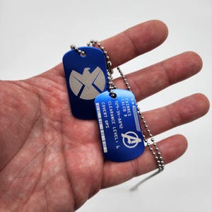 May include: Two blue metal dog tags on a silver chain. The top tag features a silver shield with a bird inside. The bottom tag has the text "FAIRRES CALEB D 729-34-8492 CLEARANCE LEVEL: 6 COVERT OPS" and a silver Avengers logo.
