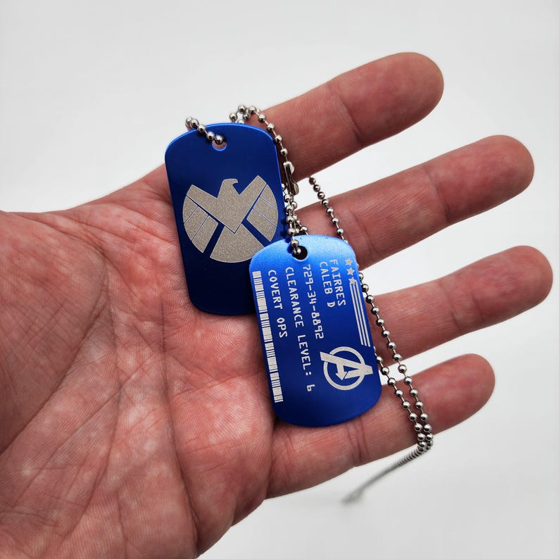 May include: Two blue metal dog tags on a silver chain. The top tag features a silver shield with a bird inside. The bottom tag has the text "FAIRRES CALEB D 729-34-8492 CLEARANCE LEVEL: 6 COVERT OPS" and a silver Avengers logo.