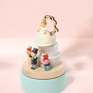 May include: A white wedding cake music box with a bride and groom figurine on top. The bride is wearing a white dress and a crown, and the groom is wearing a black suit and a top hat. The music box is decorated with a heart and a bow.