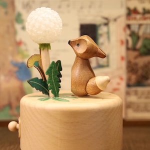 Op de afbeelding: Een houten muziekdoos met een gesneden vosbeeldje en een witte paardenbloem. De muziekdoos is gemaakt van licht hout en heeft een eenvoudig ontwerp.