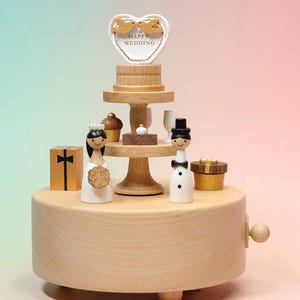 Può includere: Un carillon in legno con un supporto a più livelli e due figurine in legno, una con un abito bianco e l'altra con un completo bianco e papillon nero. In cima c'è un cartello a forma di cuore con due uccellini e la scritta HAPPY WEDDING e dei regali.