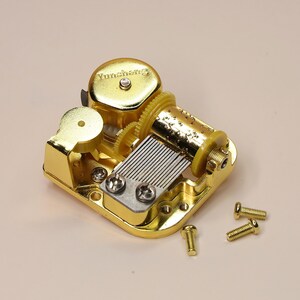 K&ouml;nnte beinhalten: Ein goldfarbener mechanischer Musikbox-Mechanismus mit sichtbaren Zahnr&auml;dern, einer Walze und einem Kamm. Auf der Musikbox ist das Wort "Yunsheng" aufgedruckt. Mehrere kleine goldene Schrauben liegen in der N&auml;he verstreut.