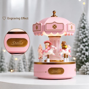 Peut inclure: Une boîte à musique carrousel rose avec un fleuron doré au sommet et un pilier central. La base et le toit sont ornés de motifs de trèfle dorés. Une plaque dorée sur la base affiche le texte gravé Stella en écriture cursive.