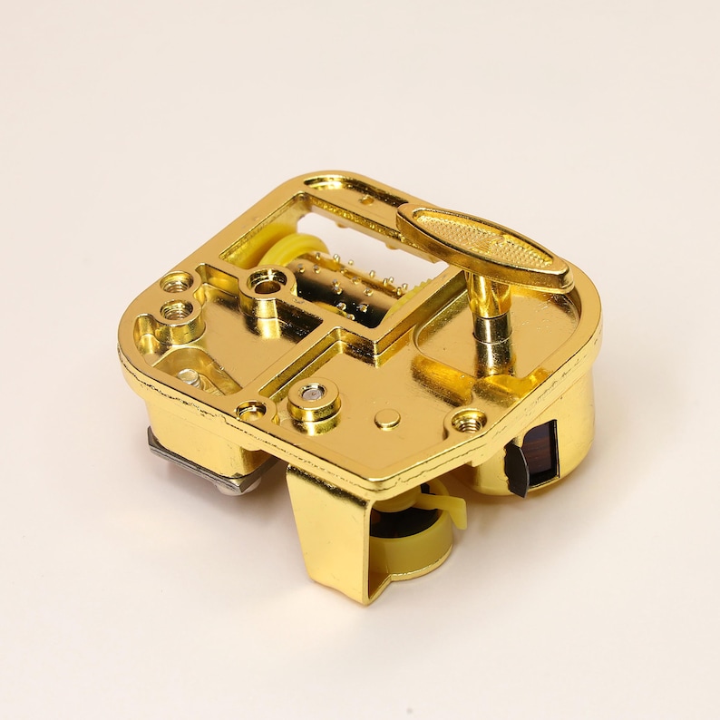 K&ouml;nnte beinhalten: Ein goldfarbener Musikbox-Mechanismus mit Handkurbel. Der Mechanismus hat eine rechteckige Form mit einer Reihe von Stiften und einem rotierenden Zylinder. Die Kurbel befindet sich oben rechts, und das Gesamtdesign ist verziert und detailliert.