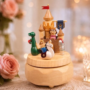 May include: A wooden music box featuring a castle with a red flag and a blue star banner. The scene includes a green dragon, a knight on a white horse, a figure in a pink dress, and a silver knight helmet, all set on a light wood circular base.