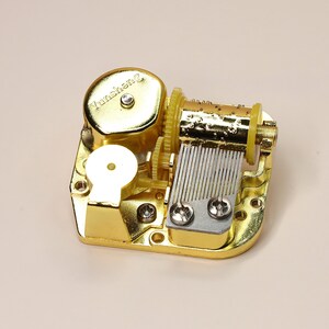 K&ouml;nnte beinhalten: Ein goldfarbener mechanischer Musikbox-Mechanismus. Der Mechanismus verf&uuml;gt &uuml;ber Zahnr&auml;der, einen Zylinder mit Stiften und einen Kamm aus Metallz&auml;hnen. Der Text "Yunsheng" ist auf der Oberseite des Mechanismus zu sehen.
