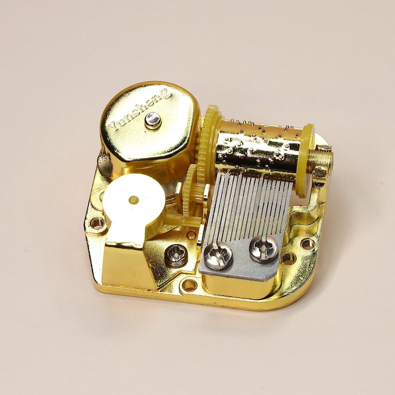 K&ouml;nnte beinhalten: Ein goldfarbener mechanischer Musikbox-Mechanismus. Der Mechanismus verf&uuml;gt &uuml;ber Zahnr&auml;der, einen Zylinder mit Stiften und einen Kamm aus Metallz&auml;hnen. Der Text "Yunsheng" ist auf der Oberseite des Mechanismus zu sehen.