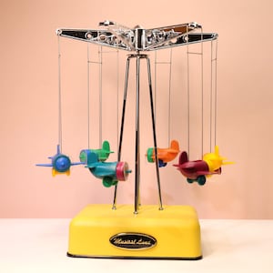 May include: A colorful, vintage-style musical carousel with five toy airplanes. The planes are in various colors, including blue, green, orange, red, and yellow. The base is yellow, and the text "Musical Land" is on the base.