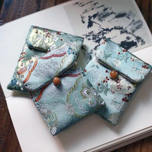 May include: Three small, blue and gold patterned fabric pouches with a flap closure and a wooden button. The pouches are made of a soft, silky material and have a traditional Chinese design.