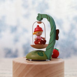 May include: A wooden music box with a green leafy design. A small wooden figure of a girl with blonde hair and a red hat sits on a swing attached to the music box. The girl is wearing a pink dress and white wings. The music box has a wooden base and a small red strawberry on the side.