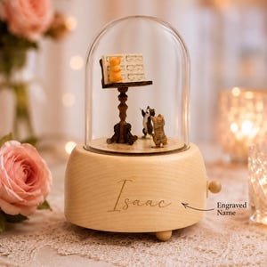 Puede incluir: Una caja de música de madera con una cúpula de cristal que contiene tres figuras de gatos en miniatura y un atril con partituras. La base es de madera clara con el nombre Isaac grabado en el frente, junto a una pequeña flecha que señala el texto.
