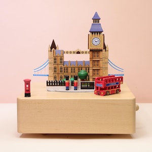 Op de afbeelding: Een houten muziekdoos met Londense bezienswaardigheden. Het bevat een miniatuur Big Ben-klokkentoren, een rode dubbeldekkerbus met de tekst "London", een rode telefooncel en speelgoedsoldaten. De doos is gemaakt van lichtgekleurd hout.