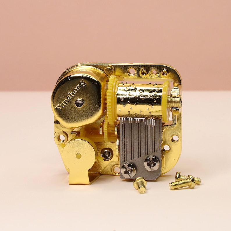 K&ouml;nnte beinhalten: Ein goldfarbenes mechanisches Spieluhrwerk mit sichtbaren Zahnr&auml;dern, einem Kamm und Schrauben. Der Text "Yunsheng" ist auf der Oberseite aufgedruckt. Die Spieluhr ist so konzipiert, dass sie eine Melodie spielt, wenn sie aufgezogen wird.