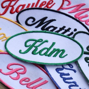 May include: A collection of embroidered name patches in various colors. Each patch features a white oval with a name embroidered in a contrasting color, such as red, black, green, and pink. The patches are on a wooden surface.