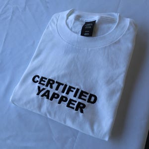 May include: White t-shirt with black embroidered text that reads "CERTIFIED YAPPER".