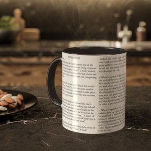 May include: Black coffee mug with a white handle. The mug features a black and white image of a page from the book of Revelation. The text on the page reads "Revelation" and includes verses from the Bible.