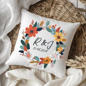 Peut inclure: Un coussin d&eacute;coratif carr&eacute; blanc orn&eacute; d'une illustration de couronne florale color&eacute;e dans des tons orange, jaune, bleu sarcelle et blanc. Au centre, les lettres R & J sont inscrites en noir au-dessus de la date 21.05.2017 en &eacute;criture cursive.