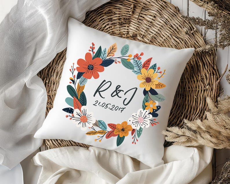 Peut inclure: Un coussin d&eacute;coratif carr&eacute; blanc orn&eacute; d'une illustration de couronne florale color&eacute;e dans des tons orange, jaune, bleu sarcelle et blanc. Au centre, les lettres R & J sont inscrites en noir au-dessus de la date 21.05.2017 en &eacute;criture cursive.