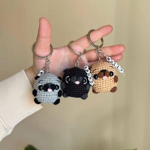May include: Three crocheted pug keychains in grey, black, and tan. Each features a metal ring, black bead eyes, a small pink tongue, and a dangling white bead charm with black letters spelling names like Bruno and Molly attached to the metal chain.