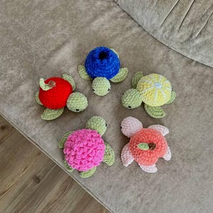 May include: Five handmade crochet turtle toys with colorful shells. The shells are red, blue, yellow, pink, and peach. The turtles have green flippers and heads. Each turtle has a unique shell design.