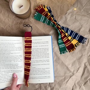 Pode incluir: Um marcador de livros de crochê feito à mão com um par de óculos pretos e uma borla. O marcador de livros é vermelho e dourado, com um padrão listrado. Vários outros cachecóis de crochê verdes, vermelhos, amarelos e azuis também são visíveis, junto com uma vela acesa e um livro aberto.