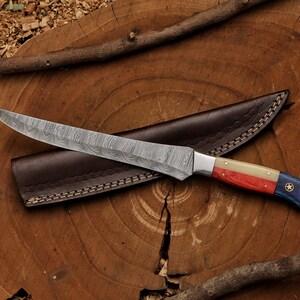 K&ouml;nnte beinhalten: Ein Jagdmesser aus Damaststahl mit einem rot-wei&szlig;-blauen Griff mit einem Stern. Das Messer befindet sich in einer braunen Lederscheide.