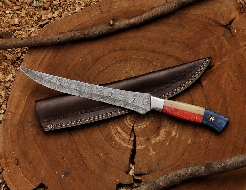 K&ouml;nnte beinhalten: Ein Jagdmesser aus Damaststahl mit einem rot-wei&szlig;-blauen Griff mit einem Stern. Das Messer befindet sich in einer braunen Lederscheide.