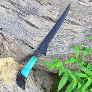 K&ouml;nnte beinhalten: Ein schwarzes und blaues Messer mit einer strukturierten Klinge und einem Holzgriff. Das Messer liegt auf einem St&uuml;ck Rinde.