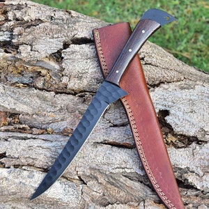 K&ouml;nnte beinhalten: Ein schwarzes Messer mit einer strukturierten Klinge und einer braunen Lederscheide. Das Messer hat einen Holzgriff mit schwarzer Oberfl&auml;che.