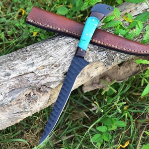 K&ouml;nnte beinhalten: Ein schwarzes und blaues Jagdmesser mit einer strukturierten Klinge und einem t&uuml;rkisfarbenen und braunen Griff. Das Messer liegt auf einer braunen Lederscheide und einem Baumstamm.