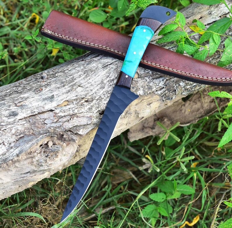 K&ouml;nnte beinhalten: Ein schwarzes und blaues Jagdmesser mit einer strukturierten Klinge und einem t&uuml;rkisfarbenen und braunen Griff. Das Messer liegt auf einer braunen Lederscheide und einem Baumstamm.