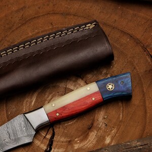 K&ouml;nnte beinhalten: Ein handgefertigtes Messer mit einer Damastmesserklinge und einem Holzgriff. Der Griff ist mit roten, wei&szlig;en und blauen Streifen verziert, die an die Flagge von Texas erinnern. Ein einziger goldener Stern ist auf dem blauen Teil des Griffs sichtbar.