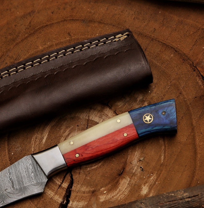K&ouml;nnte beinhalten: Ein handgefertigtes Messer mit einer Damastmesserklinge und einem Holzgriff. Der Griff ist mit roten, wei&szlig;en und blauen Streifen verziert, die an die Flagge von Texas erinnern. Ein einziger goldener Stern ist auf dem blauen Teil des Griffs sichtbar.