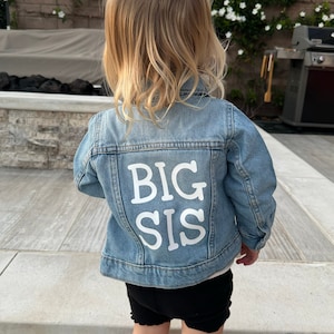 Może przedstawiać: Małe dziecko w niebieskiej dżinsowej kurtce z napisem "BIG SIS" w białych literach na plecach. Dziecko ma na sobie czarne spodenki i związane włosy w kok.