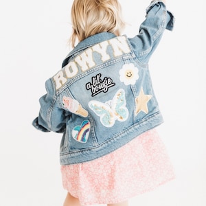 May include: A blue denim jacket with a white embroidered name "ROWYN" on the back. The jacket has several colorful patches, including a butterfly, a rainbow heart, a smiley face, a star, and an ice cream cone. The jacket also has a black patch with the text "a lil boujie".