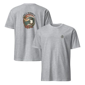 May include: A heather grey t-shirt with a graphic of a surfer on a wave with the text "Big Z Memorial Surf Off" on the back. The front of the shirt has a small embroidered logo of a "Z" in a circle.