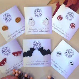 May include: Six pairs of stud earrings on white display cards featuring seasonal motifs: orange pumpkins, white ghosts, red and white mushrooms, red vampire lips with fangs, black bats, and white skulls with black eyes. Each card includes SMR designs branding.