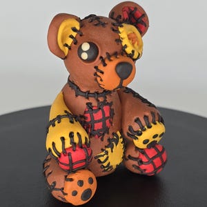 May include: A brown teddy bear figurine with orange, yellow, and red details. The bear features black stitching and patches, with a black nose and button eyes. The bear is sitting on a black surface.