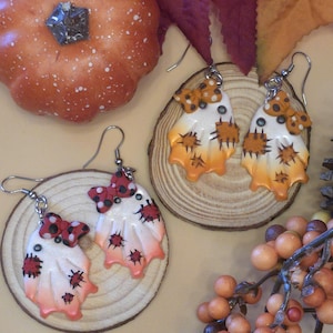 May include: Two pairs of ghost-shaped earrings featuring white bodies with orange or red gradient bottoms, small bows, and stitched patch details. The earrings have silver-toned metal hooks and are displayed on circular wooden slices near a pumpkin.