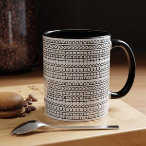 Könnte beinhalten: Eine weiße Keramiktasse mit schwarzem Innenraum und Griff. Die Tasse ist mit einem sich wiederholenden schwarzen geometrischen Muster verziert. Ein silberner Löffel und ein Macaron liegen auf einer Holzoberfläche.