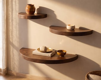 May include: Three semi-circular dark wood floating shelves mounted on a wall. The top shelf holds a brown ceramic jar. The middle shelf holds a small white candle. The bottom shelf holds a book, a gold-toned dish with a stone, and a small bowl.