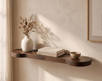 May include: A dark brown wooden wall shelf with rounded edges holding a white ceramic vase filled with dried flowers, a stack of two beige books, and a small beige ceramic bowl. The shelf is mounted horizontally on a light beige wall.